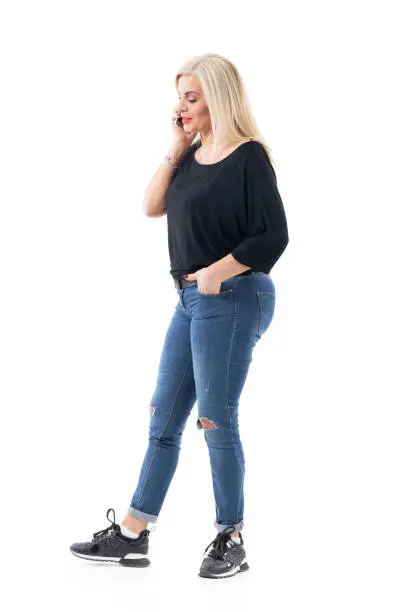 Side view of serious confident blonde casual middle age woman walking and talking on mobile phone. Full body length isolated on white background.