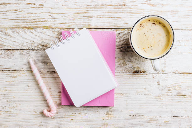 istock Home office desk, plan writing or sketching concept. Open notebook with blank page, cup of coffee and donut covered with pink icing on a white rustic wooden background. Top view. Flat lay 1199320290