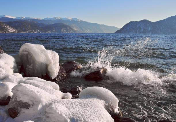 Ice and snow on the rocks on the shore of lake Teletskoye, which does not freeze in winter. Waves, mountains, blue sky in the distance. Without people-the nature of Siberia. Russia, Gorny Altai, 2020