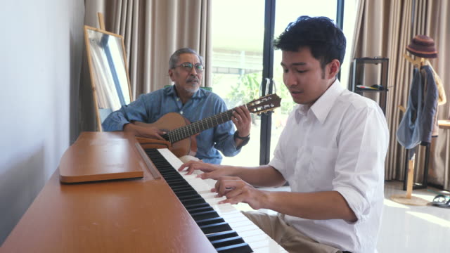 Asian adult son enjoy playing piano and senior father playing guitar together in living room at home