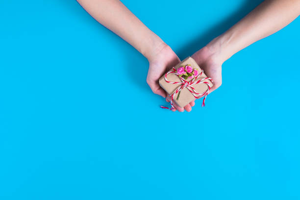 istock both hands holding on a small brown gift box tied with a red and white rope, that decorated with a few small pink roses, those placed on blue background 1199377797