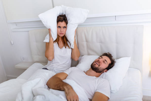 les jeunes couples ont des problèmes avec le ronflement de l'homme. couples hétérosexuels dans le lit, l'homme dort et ronfle avec la bouche ouverte, tandis qu'une femme fatiguée irritée par le ronflement s'asseyant sur le lit avec un oreiller sur sa  - mouth open women displeased anger photos et images de collection