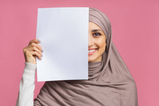 lächelnde arabische mädchen bedeckt die hälfte des gesichts mit leeren papierblatt - veil human face women fashion model stock-fotos und bilder