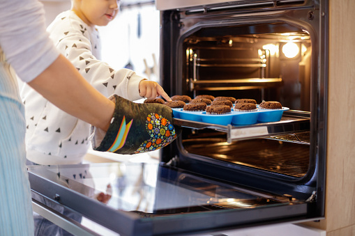 Mother taking baked muffins out of oven while her son is trying to touch one