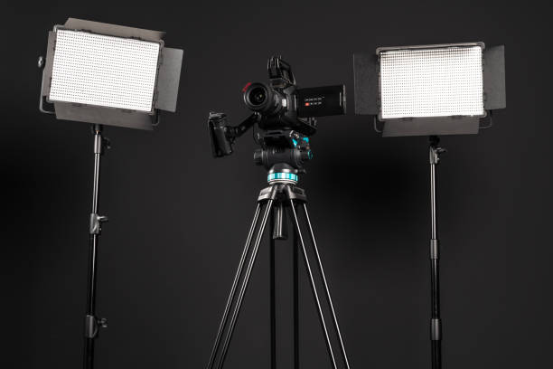 istock Professional cinema video camera on a camera tripod and one lighting LED panel on black studio background, low angle view 1199401510