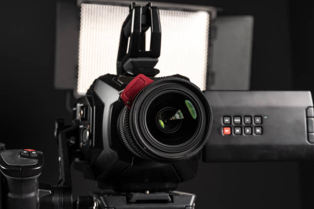 istock Professional cinema video camera on a camera tripod and one lighting LED panel on black background, close-up 1199401545