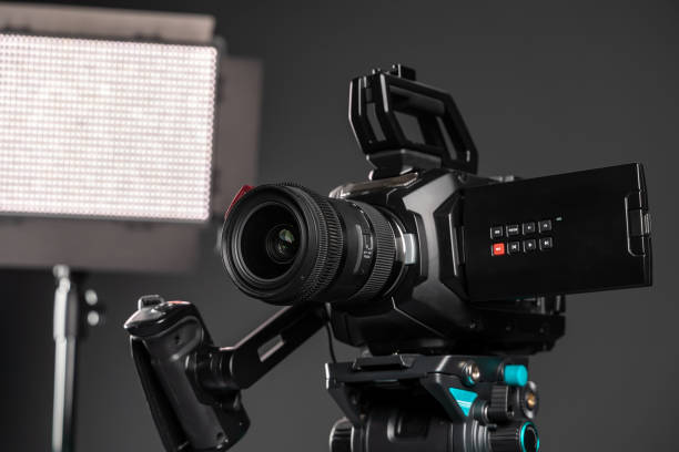 istock Professional cinema video camera on a camera tripod and one lighting LED panel on black studio background, close-up 1199402393