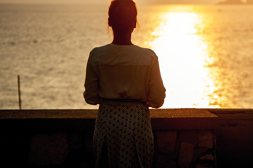 Young woman standing looking at sunset in front of the sea on Ponza island coast, on a wall with view of the ocean. 9843