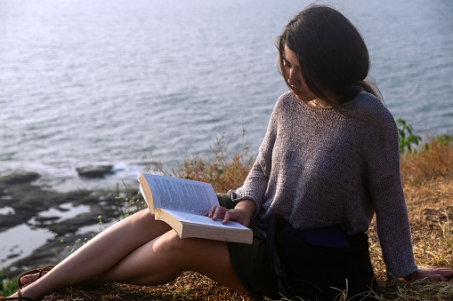 Attractive Asian woman is sitting on the edge of the mountain with a sea view, reading a book. Close-up portrait