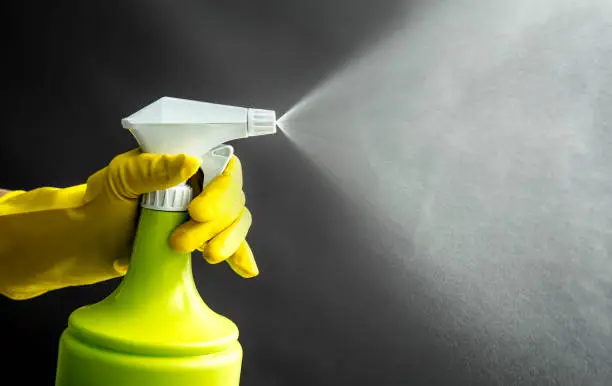 Photo of Woman wearing yellow rubber gloves using green spray bottle and spraying liquid mist in air, cool lighting effect. Lot of copy space.
