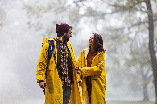 Happy couple in yellow raincoats walking and communicating during rainy day in misty nature. Copy space.