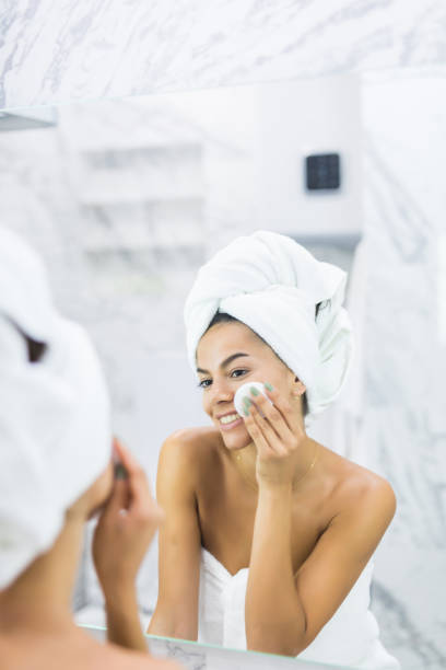 istock Beautiful young woman touching her face with sponge and smiling while standing in front of the mirror in bathroom 1199415109