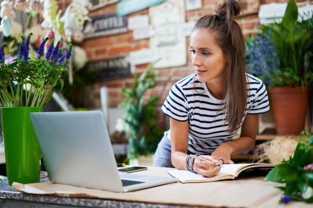 istock Young florist writing in notebook and looking at laptop while leaning on counter 1199418350