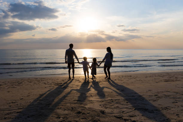 happy asian family on summer vacation father, mother, son and daughter holding hands running down to the seashore together. background sunset in sea. relax holiday and travel concept. - asian ethnicity child four people couple imagens e fotografias de stock