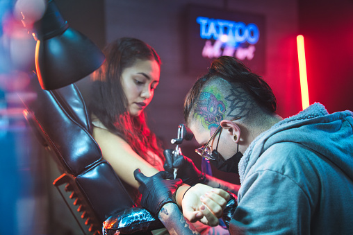 Young people in tattoo studio. Person in beauty parlor and fashion salon for modern tattooing and body art. Latino female teenager getting tattoos on arm skin by professional tattoist