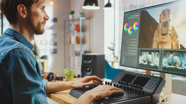 istock Male Video Editor with Beard and Jeans Shirt Works with Footage on His Computer Using an Editing Console Deck. He Works in a Cool Office Loft. Other Female Creative Colleague Works in the Background. 1199435105
