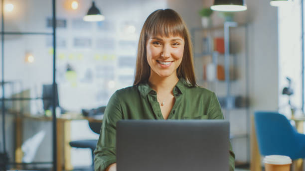 istock Young Beautiful Brunette Works on a Laptop Computer in Cool Creative Agency in a Bright Loft Office. She Smiles and Laughs on Camera. 1199435107