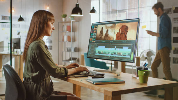 istock Beautiful Brunette Female Video Editor Works with Footage on Her Personal Computer with Big Display. She Works in a Cool Office Loft. Other Male Creative Colleague Walks in the Background. 1199435110