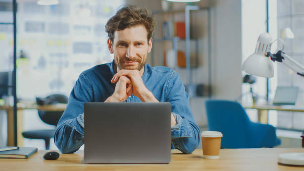 istock Young Handsome Man Works on a Laptop Computer in Cool Creative Agency in a Loft Office. He has a Take-away Coffee and a Notebook on the Table. He Wears a Jeans Shirt. 1199435120