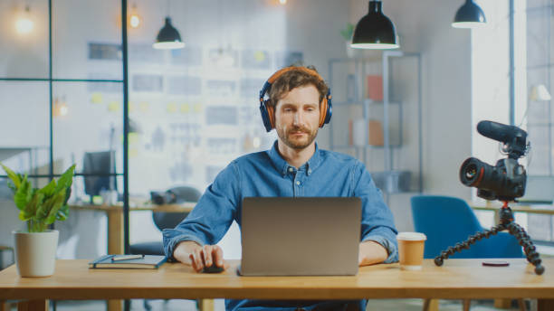 istock Young Creative Blogger Works on a Laptop Computer in Cool Creative Agency in a Loft Office. He Editing a New Video or Photos. He Wears a Jeans Shirt and Headphones. 1199435155