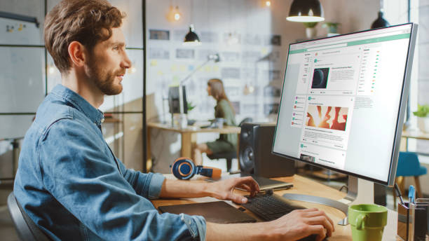 istock Beautiful Male Scrolls Through Social Media Site on His Personal Computer with Big Display. He Works in a Cool Office Loft. Other Female Creative Colleague Works in the Background. 1199435184
