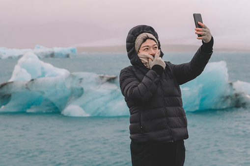 an asian chinese female traveler in front of iceberg glacier of iceland during overcast day with warm clothing hooded shirt