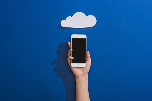 cropped view of woman holding smartphone near empty white paper cloud on blue