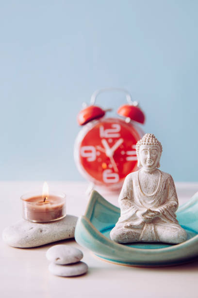 Finding time for self care and balance between body soul and mind. Balance between work and home concept. Lotus pose white color sitting meditation Buddha figurine, red alarm clock on background.