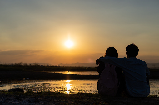Romantic couple in love sitting together at sunset lake, silhouettes of young man and woman on holidays or honeymoon