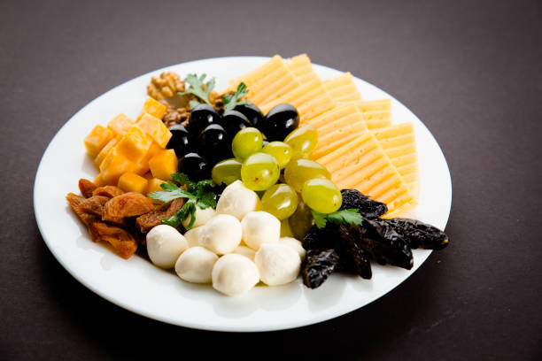 Cheese plate for wine tasting - cheddar cheese, monterey cheese, colby jack cheese, mozzarella cheese balls, fried figs and apricots, fresh grapes, black olives and walnuts on white plate on black background