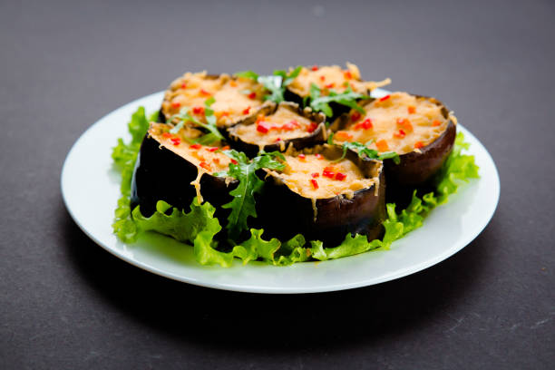 Roasted eggplants halves stuffed with tomatoes, sweet peppers and rise, covered with shredded cheddar cheese served on white plate with greens