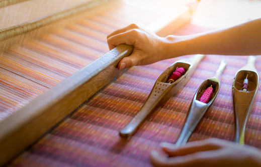 Woman working on weaving machine for weave handmade fabric. Textile weaving. Weaving using traditional hand weaving loom on cotton strands. Textile or cloth production in Thailand. Asian culture.