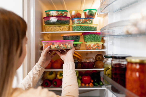 Woman taking raw food from refrigerator Close up shot of woman taking container with frozen mixed vegetables from refrigerator while looking at camera. storage compartment stock pictures, royalty-free photos & images