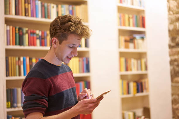 istock Young student in library uses his phone in front of bookshelf 1199468236