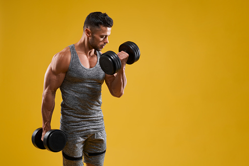 Side view of handsome athletic bodybuilder lifting heavy weight. Strong brutal guy in sportswear pumping up muscles. Isolated on yellow studio background with copy space. Concept of sport.