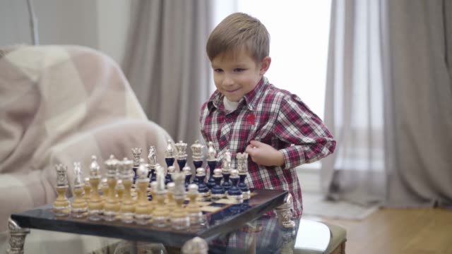 Portrait of smart Caucasian boy setting chess pieces on board. Little cute child starting playing with unrecognizable father. Intellect, intelligence.