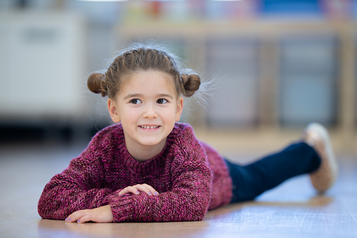 A beautiful little Caucasian girl with buns in her hair, lays on her Kindergarten classroom floor. She is dressed in a casual in denim pants and a sweater and smiling as she poses for the portrait.