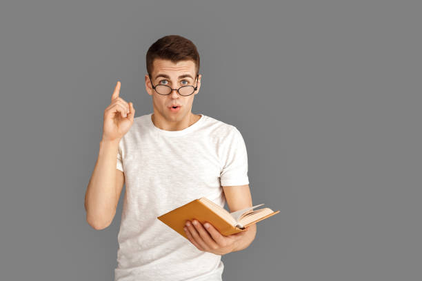 istock Freestyle. Young student in eyeglasses standing isolated on grey with book pointing up idea 1199506965