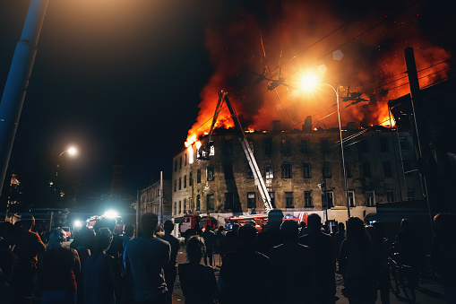 Burning city building at night, Firefighters extinguish house, Silhouettes of citizens people on foreground looks and flaming fire with smoke.