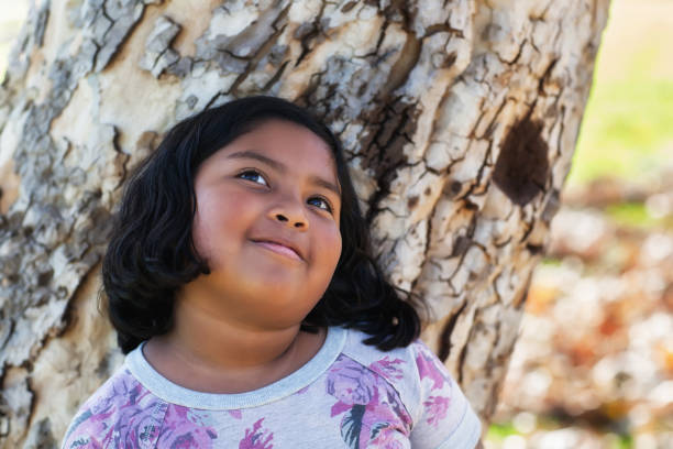 A 9 year old girl daydreaming while leaning on a tree trunk.