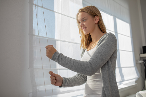 Pretty, young woman lowering the interior shades/blinds in her modern interior apartment