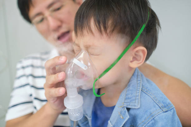 Asian father helping his toddler son with inhalation therapy by the mask of inhaler. Sick little kid with respiratory problem with oxygen mask breathes through nebulizer Asian father helping his toddler son with inhalation therapy by the mask of inhaler. Sick little kid with respiratory problem with oxygen mask breathes through nebulizer at doctor clinic pediatric nebulizer mask stock pictures, royalty-free photos & images