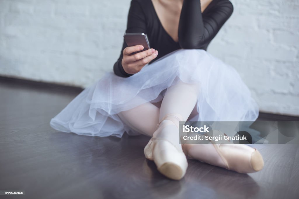 Young ballet dancer sitting leaning white wall using phone in studio active lifestyle close-up Young female ballerina practice in studio sitting leaning white brick wall browsing smartphone close-up Smart Phone Stock Photo
