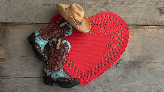 cowboy boots and hat laying on a red heart paper doily on a rustic wood background with copy space