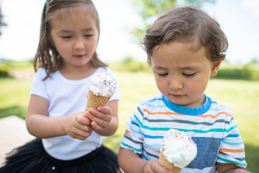 A young Asian brother and sister sit outside on a warm summer day each holding an ice cream cone that they have just been given.  They are eyeing up their treats and getting a god look at them before taking the first lick.