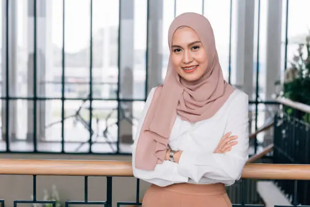 Young happy and successful South East Asian Islamic business woman with arms crossed in business corporate building setting looks at camera. She wears head scarf or hijab and modern elegant clothes