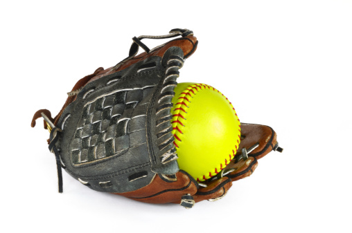 A close-up of a used yellow softball in a leather glove along with a pair of black cleats sitting in the grass