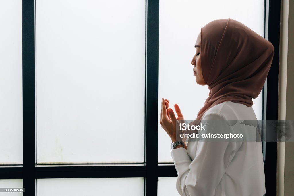 Islamic woman praying near window Southeast Asian Islamic female in hijab clasping hands and looking up while standing near window during prayer Praying Stock Photo