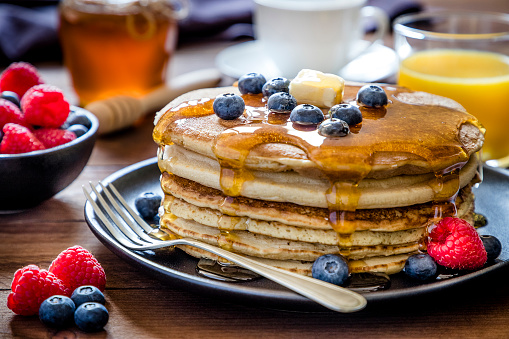 Front view of a stack of pancakes with blueberries and butter on top pouring honey on a black plate surrounded by a glass full os orange juice, a cup of coffee, a bowl with berries and a honey jar. Low key DSLR photo taken with Canon EOS 6D Mark II and Canon EF 24-105 mm f/4L