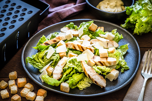 Close up of a fresh homemade chicken salad on a black plate surrounded by a fork, a napkin, croutons, a bowl with the dressing for seasoning the salad and a lettuce. The plate is at the center of the image. \nLow key DSLR photo taken with Canon EOS 6D Mark II and Canon EF 24-105 mm f/4L